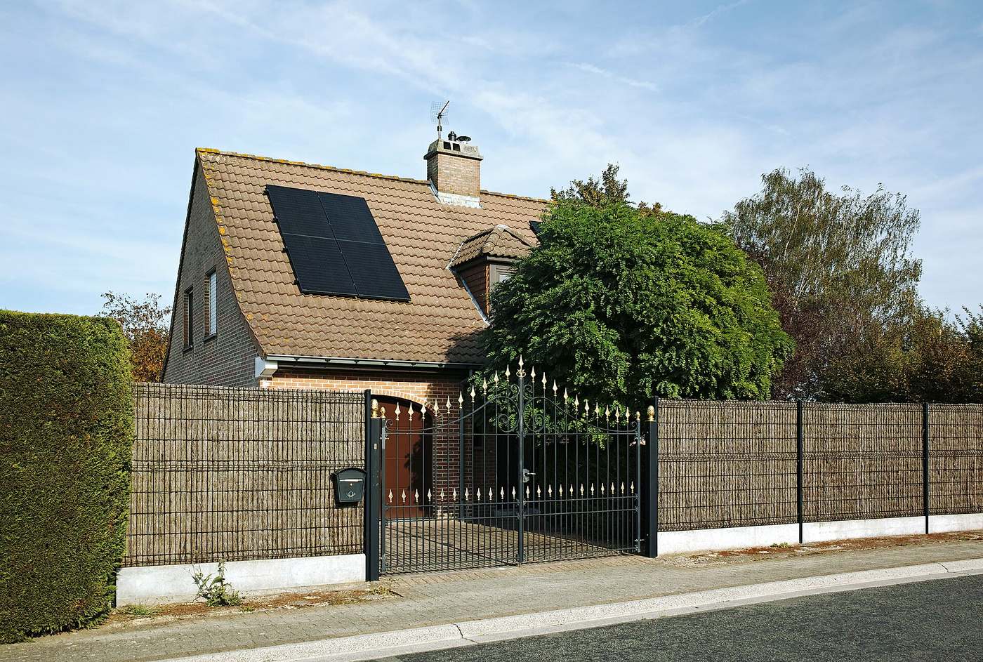 Woning met bruin pannendak en twee zwarte zonnepanelen, achter een haag, een omheining en een smeedijzeren poort